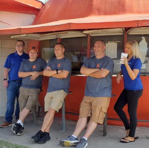 Five people stand and sit outside a retro ice cream shop, smiling and chatting, wearing casual shirts and shorts.