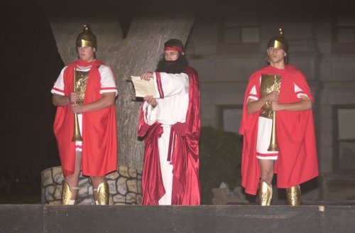 Three individuals dressed in Roman-style costumes, with one holding a scroll, standing on a stage at night.
