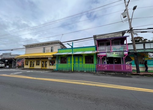 Colorful storefronts line a street, featuring yellow, green, and pink buildings under a cloudy sky.