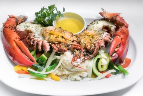 A gourmet lobster dish with stuffed lobster tails, vegetables, and a small bowl of olive oil on a white plate.