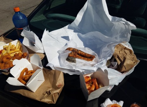 A variety of takeout food including fries, shrimp, and fish, arranged on a car hood with a drink nearby.