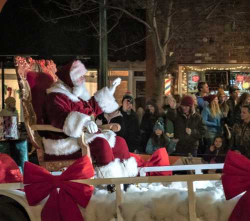 Santa Claus waves from a festive float, surrounded by a cheerful crowd during a holiday parade.