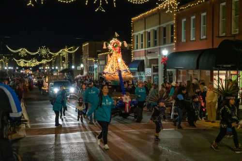 A festive night parade with people in teal shirts, decorated floats, and holiday lights lining the streets.