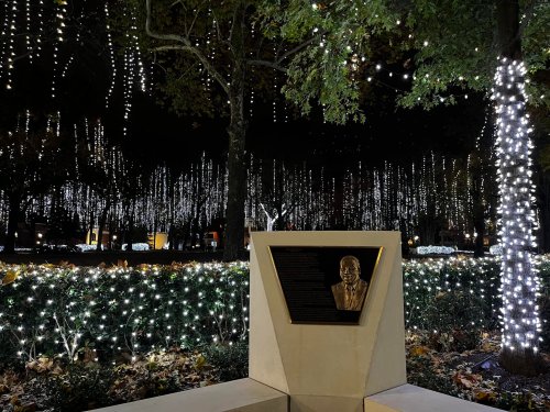 A bronze bust in a lit park, surrounded by trees adorned with sparkling white lights.