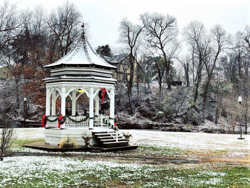 A snow-covered gazebo decorated with red ribbons, surrounded by a winter landscape and bare trees.