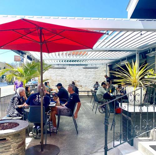 Outdoor dining area with red umbrellas, people seated at tables, and a sunny atmosphere. Green plants add a lively touch.