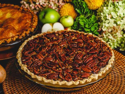 A pecan pie on a woven mat, surrounded by flowers, apples, and another pie in the background.