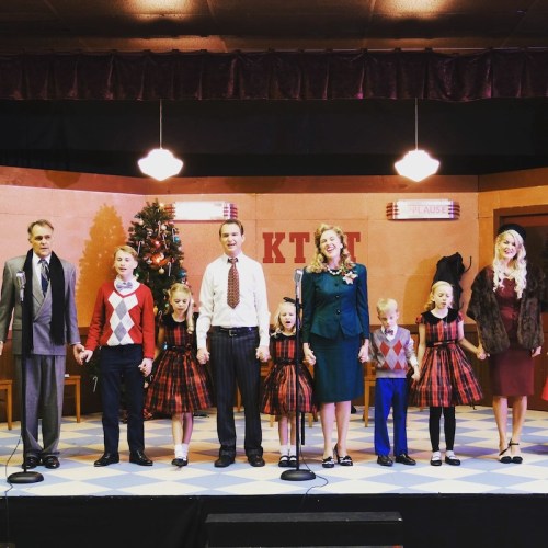 A group of children and adults in festive attire stand on stage, holding hands, with a Christmas tree in the background.