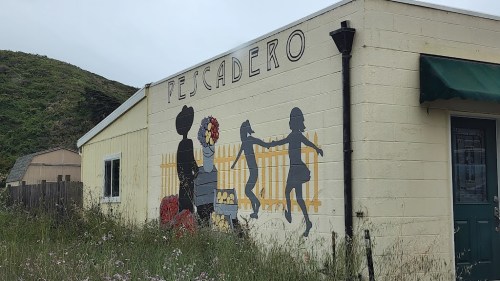Mural on a building in Pescadero featuring a woman with flowers and children playing near a fence. Green hills in background.