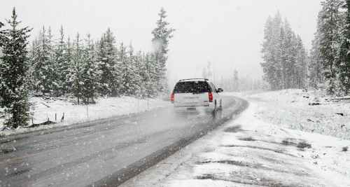A white van drives on a snowy road surrounded by evergreen trees in a winter landscape. Snow is falling heavily.