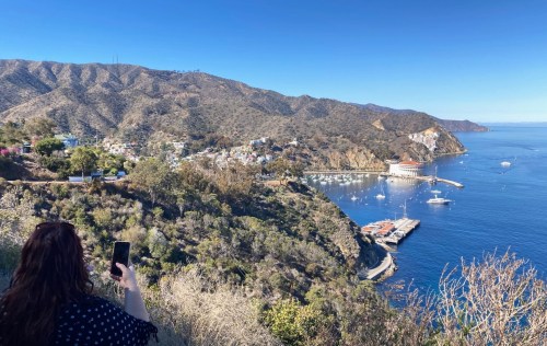 A person takes a photo of a coastal view with hills, a marina, and clear blue water on a sunny day.