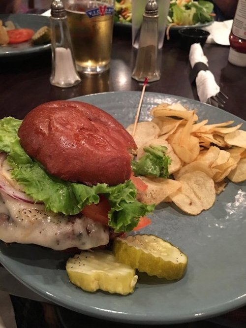 A juicy burger with lettuce, tomato, and cheese, served with potato chips and pickles on a blue plate.