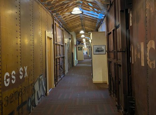 A long hallway with wooden beams, vintage walls, and overhead lights, featuring framed pictures along the sides.