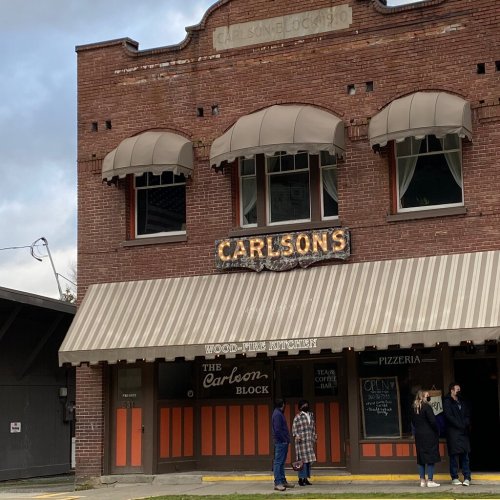 Historic brick building with "Carlsons" sign, featuring a striped awning and people standing outside a pizzeria.