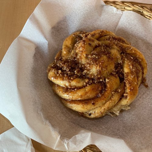 A freshly baked cinnamon roll topped with a crunchy pecan streusel, served in a paper-lined basket.