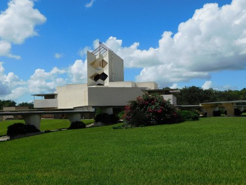 A modern architectural building with a unique design, surrounded by green grass and colorful flowers under a blue sky.