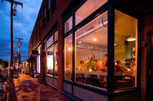 A cozy restaurant with warm lighting, filled with diners, seen from the street at dusk.