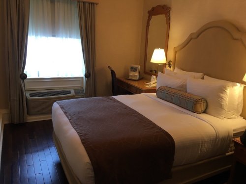 Cozy hotel room featuring a queen bed, desk, and window with sheer curtains. Warm lighting creates a welcoming atmosphere.