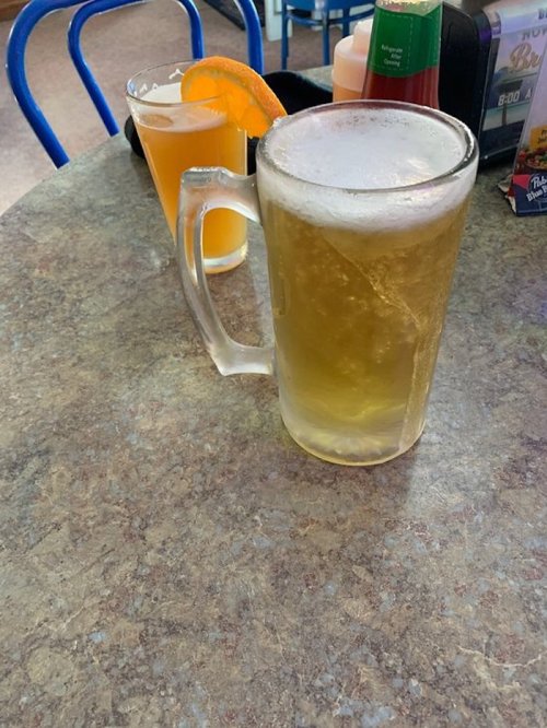 A frosty mug of beer sits on a table next to a glass of orange drink with a slice of orange.