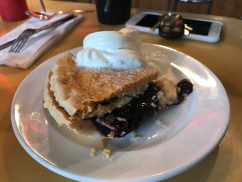 A slice of berry pie topped with whipped cream, served on a white plate with a fork and napkin in the background.