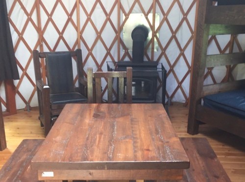 Interior of a yurt featuring a wooden table, chairs, a bunk bed, and a stove against a lattice wall.