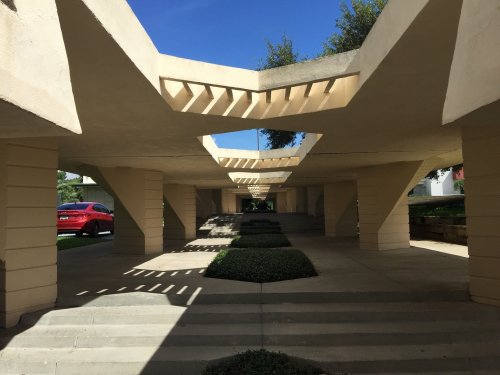 A modern architectural structure with angular columns and open spaces, featuring greenery and a blue sky above.