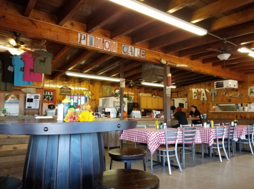 Interior of a rustic diner with wooden beams, checkered tablecloths, and a bar area. Signs read "Pinon Cafe" and "EAT."