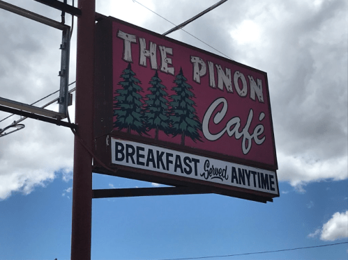 Sign for The Pinon Café featuring trees and the text "Breakfast Served Anytime" against a blue sky.