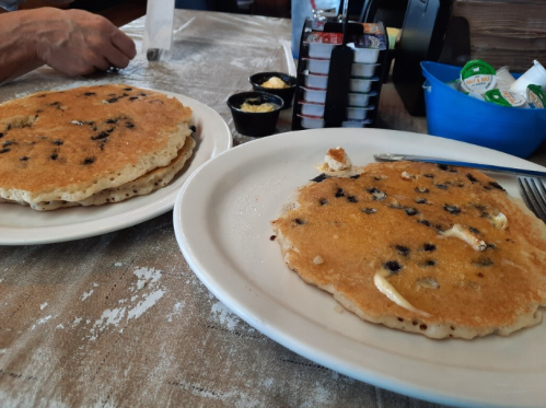 Two large pancakes with chocolate chips, one partially eaten, served with butter and small containers of syrup.