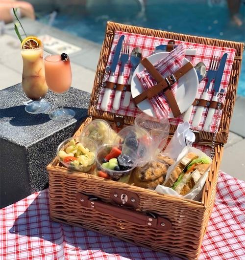 A picnic basket filled with food, next to two colorful cocktails by a poolside.