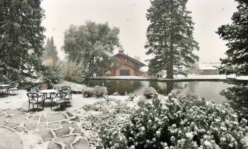 A snowy landscape featuring a pond, trees, and a rustic building, with a stone patio and chairs in the foreground.