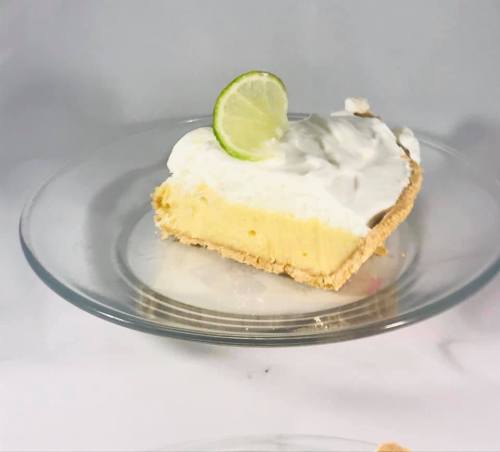 A slice of key lime pie topped with whipped cream and a lime slice, served on a clear glass plate.