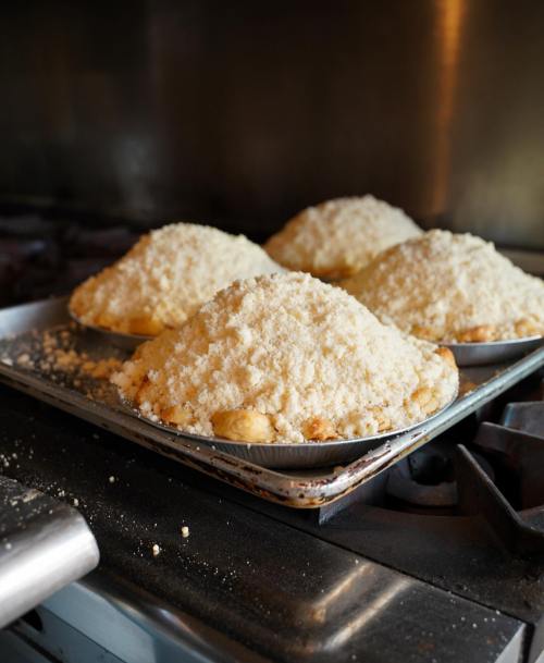Four freshly baked pies with crumbly topping on a baking tray, resting on a stove in a kitchen.
