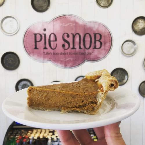 A slice of pumpkin pie on a plate, held in front of a "Pie Snob" sign with a playful slogan.