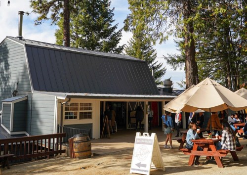 A rustic building surrounded by trees, with people dining at outdoor tables under large umbrellas.