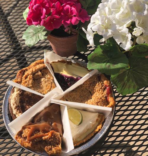 A platter of five pie slices, including fruit and cream varieties, surrounded by colorful flowers in pots.