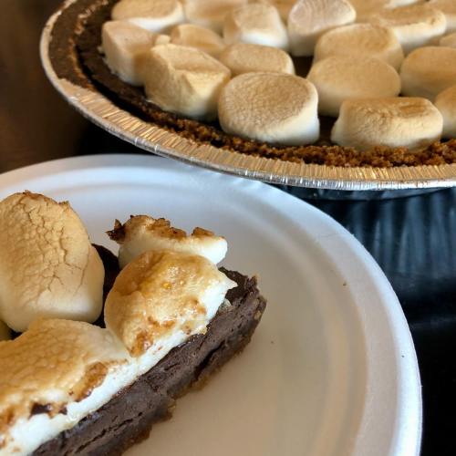A slice of chocolate pie with toasted marshmallows on a plate, next to a whole pie with more marshmallows on top.