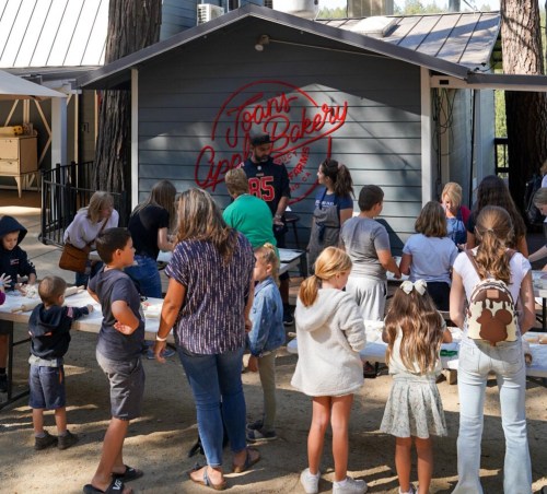 A group of people, including children and adults, gather around tables outside a bakery in a wooded area.