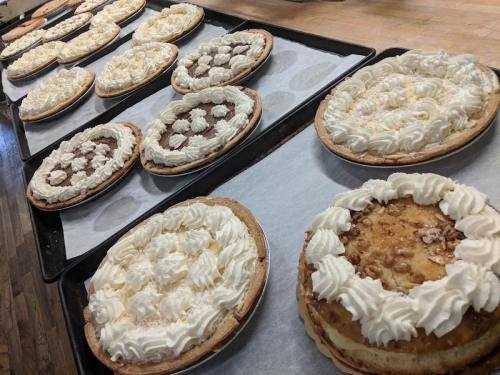 A variety of pies with whipped cream topping arranged on baking trays, showcasing different flavors and designs.