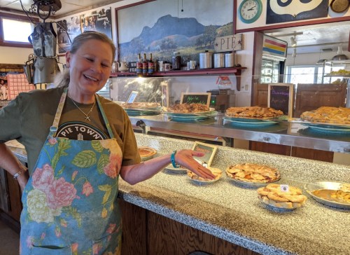 A smiling woman in an apron stands by a display of various pies in a cozy bakery setting.