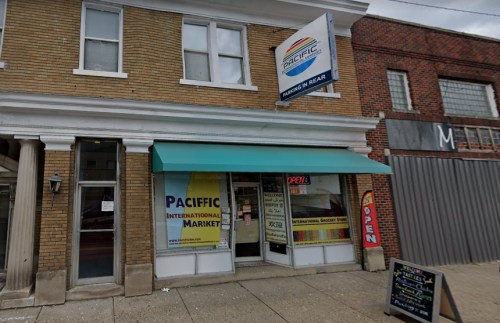 Exterior of Pacific International Market, featuring a green awning and an "Open" sign, with a brick building nearby.
