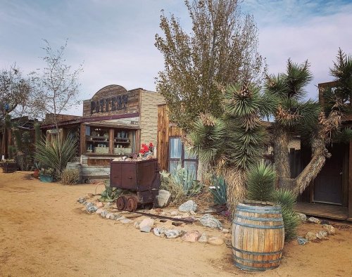 A rustic western town scene with wooden buildings, desert plants, and a vintage cart surrounded by rocks and a barrel.