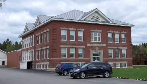 Historic red brick school building with large windows and a metal roof, surrounded by parked cars and green grass.