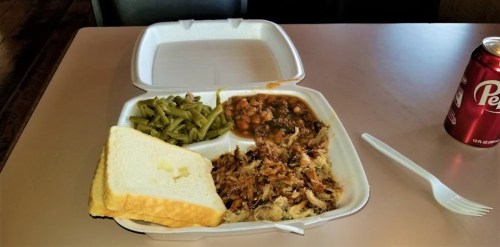 A takeout container with pulled pork, green beans, baked beans, a slice of bread, and a can of soda on a table.