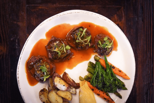 A plate featuring three meat patties in sauce, served with roasted potatoes and colorful vegetables.