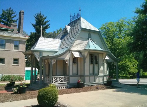 A charming, Victorian-style gazebo with a peaked roof and decorative details, surrounded by greenery and blue skies.
