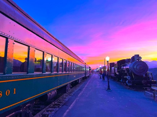 A vibrant sunset casts colorful reflections on vintage train cars at a station, with silhouettes of people and a steam engine.