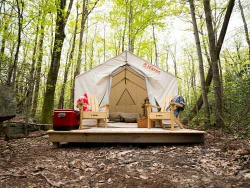 A cozy tent on a wooden deck in a forest, surrounded by trees, with two chairs and a cooler nearby.