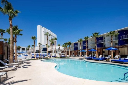 A sunny pool area surrounded by palm trees and lounge chairs, with a hotel in the background under a clear blue sky.
