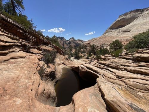 A scenic view of a rocky canyon with a narrow waterway, surrounded by trees and mountains under a clear blue sky.